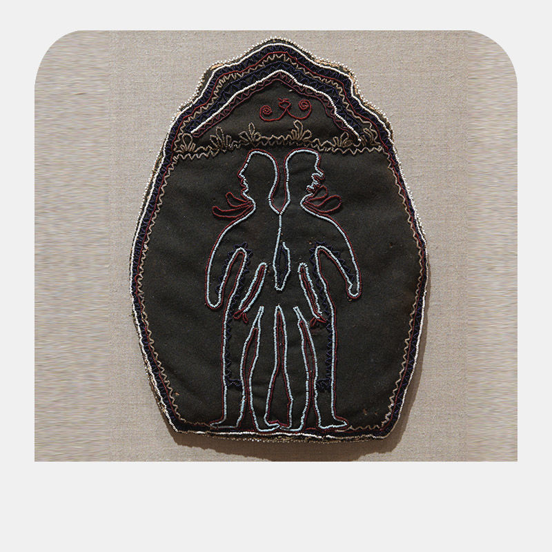 A black circular patch is woven unto pale fabric and etches a white outline of two human silhouettes.